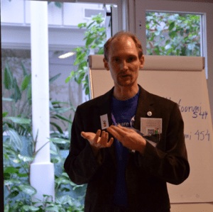 Mies seisoo puhumassa. Taustalla kasveja ja fläppitaulu.