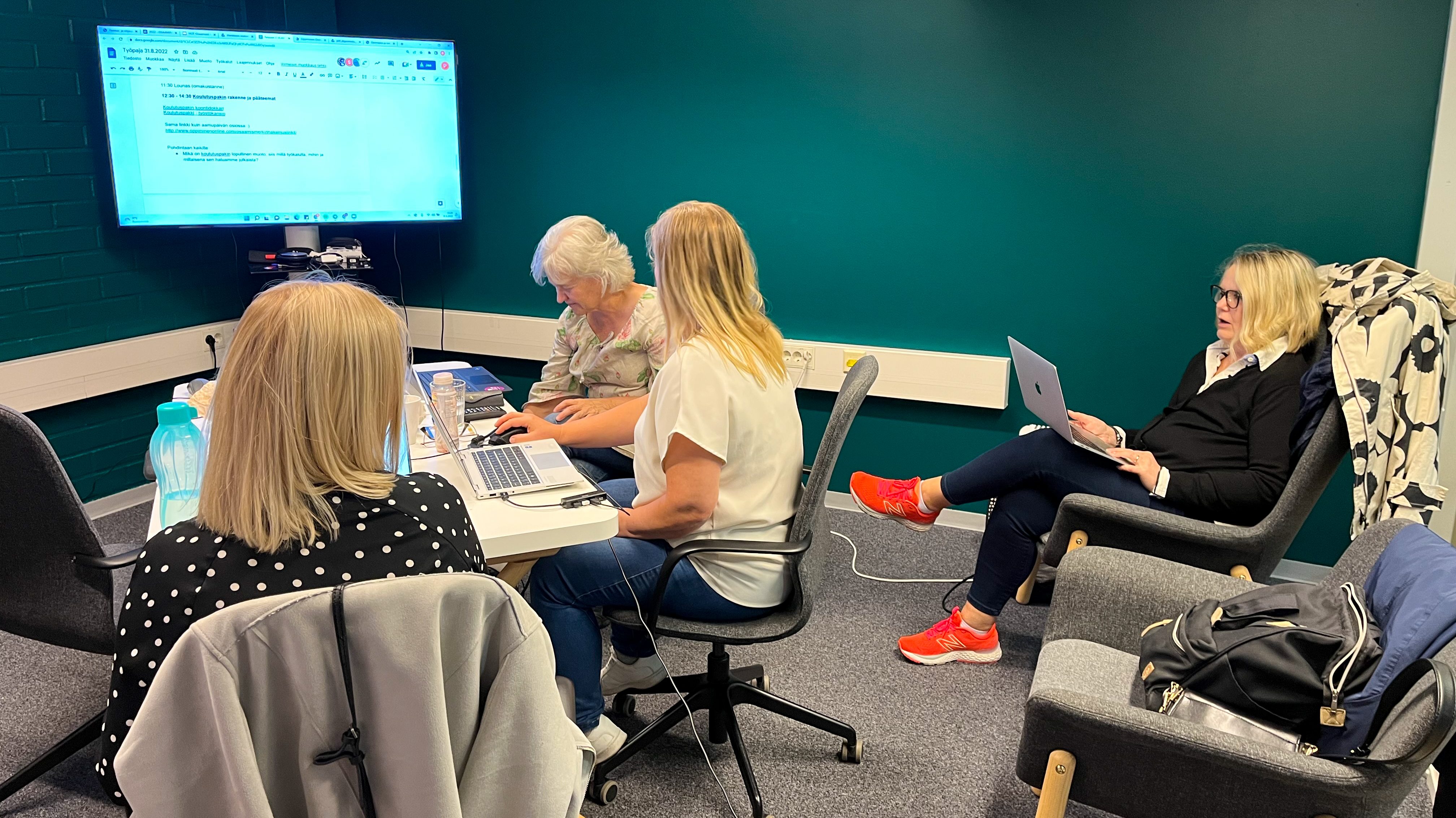 Neljä henkilöä tietokoneilla kokoushuoneessa. Taustalla iso tietokoneruutu.