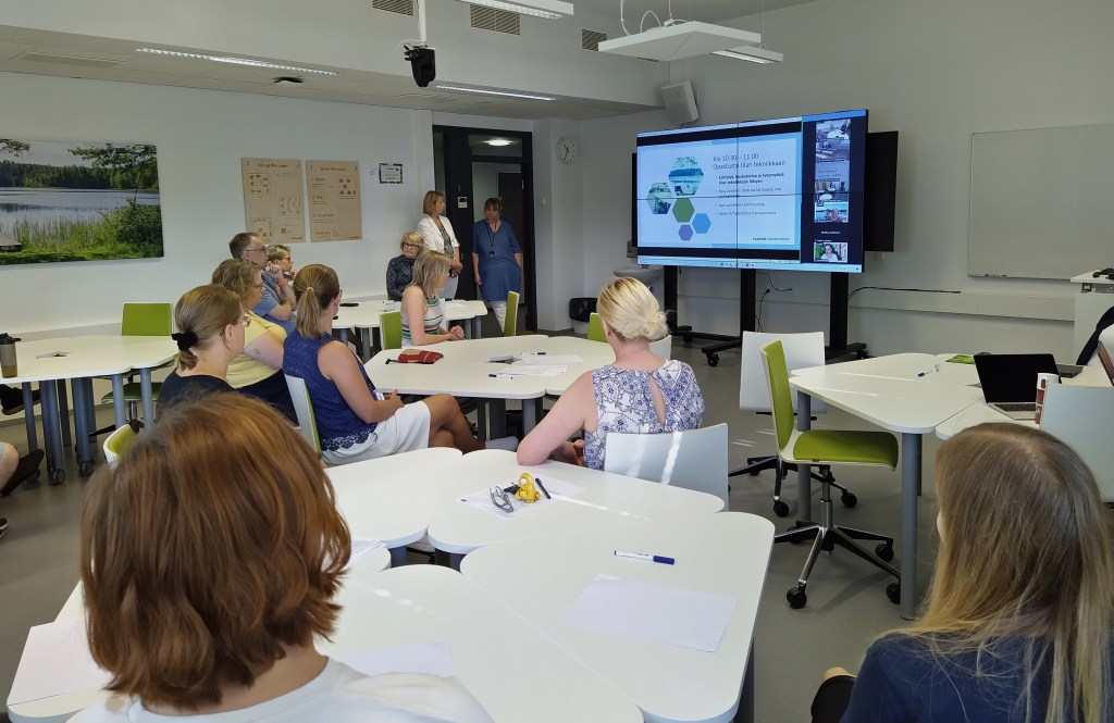 Ihmisiä istumassa luokkahuoneessa selkä kameraan. Takaseinällä iso näyttö, jossa on diaesitys menossa.