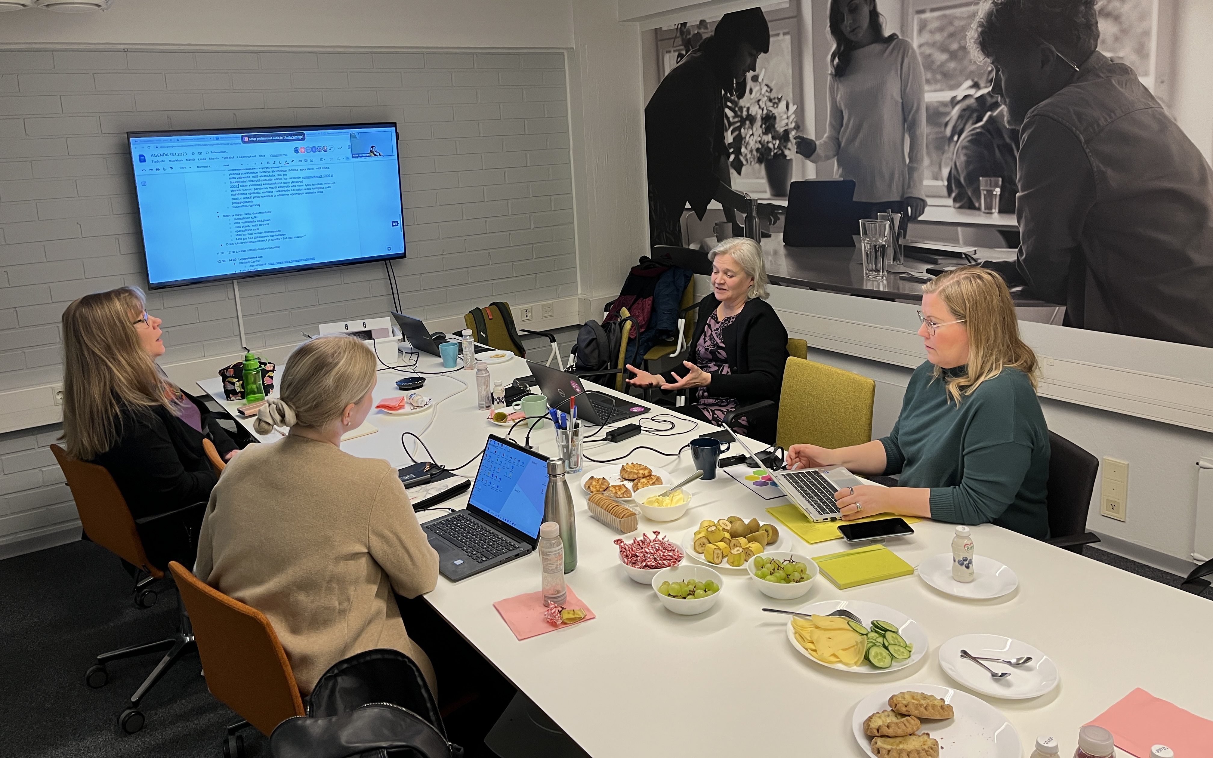 Kokoushuone. Taulutelevisio. Pöydällä tietokoneita. Neljä naista. Aamupalatarjoilu.