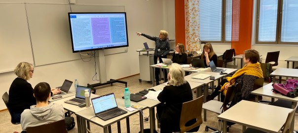 Luokkahuone. Kuusi ihmistä istuvat tietokoneiden äärellä ja katsovat, kun yksi esittää suurelta näytöltä.