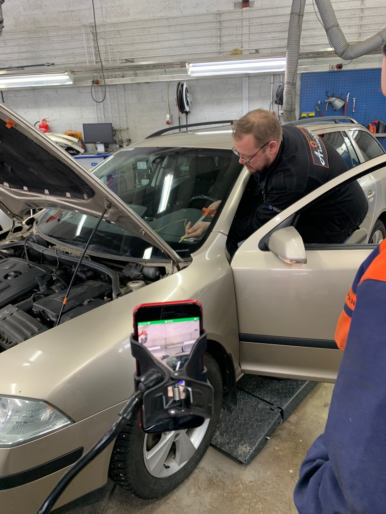 Telineessä oleva kännykkä kuvaa miestä, joka korjaa auton kojelaudalta jotain. Auton nokkapelti ja kuskin ovi ovat auki.