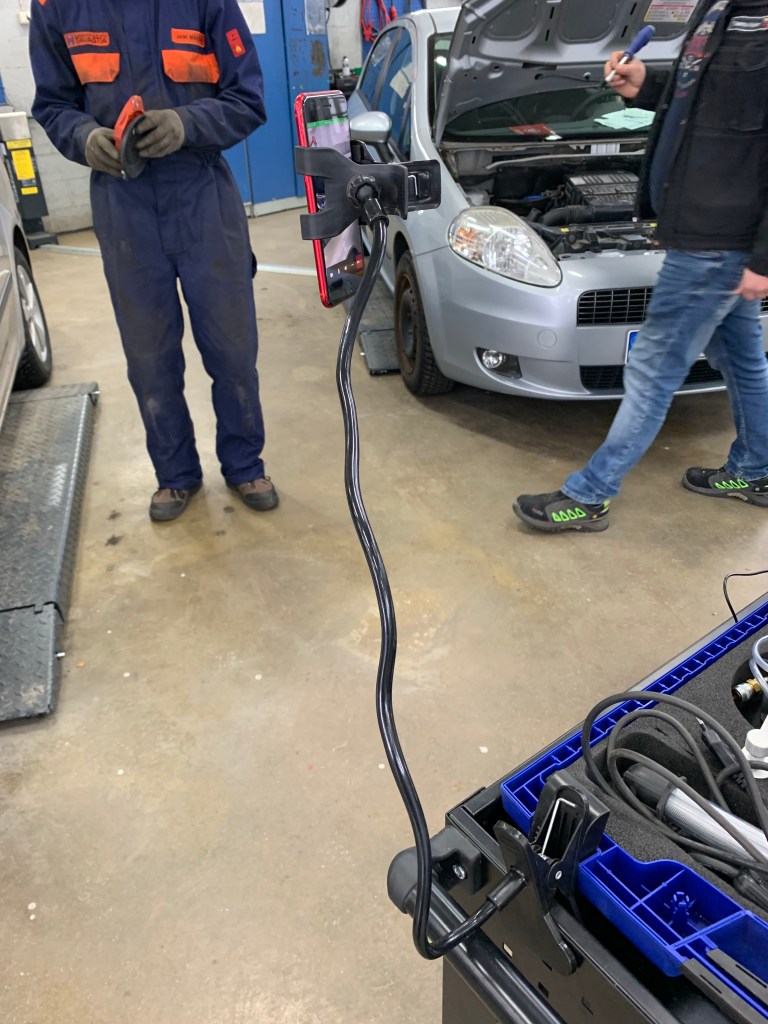 Kännykkä kuvaa pöytään kiinnitetyn telineen varassa. Taustalla auto nokkapelti auki. Ihminen seisoo työhaalarit päällä taustalla. Toinen ihminen kävelee haalari-ihmistä kohden ruuvimeisseli kädessä.