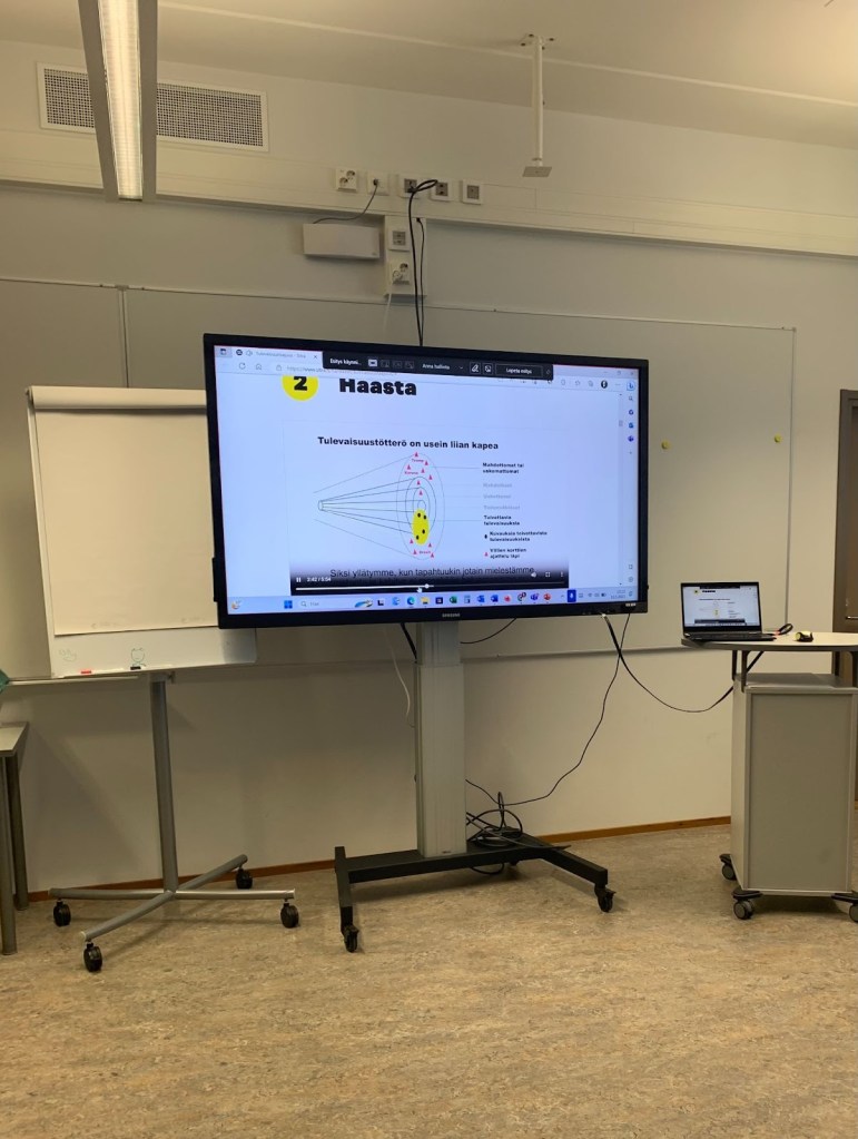 Luokkahuoneen etuosa. Siirreltävä iso televisionäyttö. Näytöllä esitys, jossa lukee Haasta.